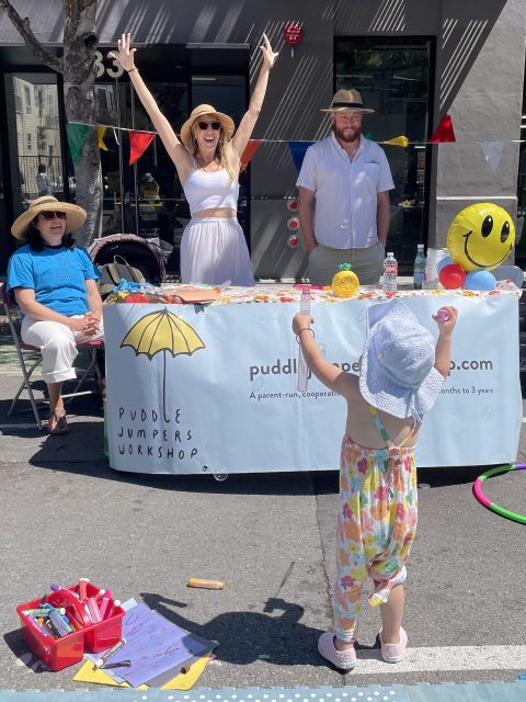 A child in a colorful jumpsuit and hat faces three adults at a booth with a "Puddle Jumpers Workshop" banner. One woman stands with arms raised, while another woman and a man sit beside the booth.