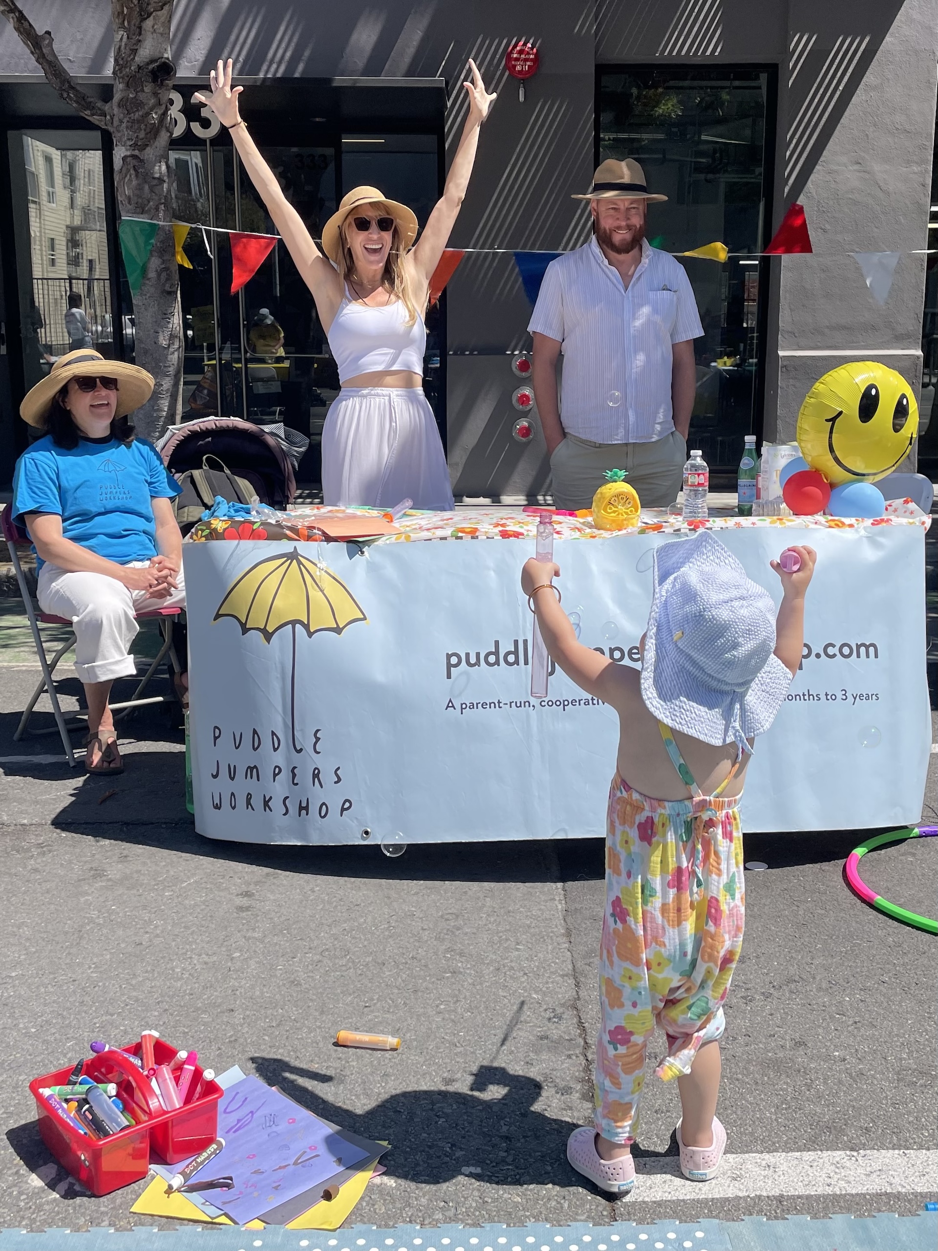 A child in a colorful jumpsuit and hat faces three adults at a booth with a "Puddle Jumpers Workshop" banner. One woman stands with arms raised, while another woman and a man sit beside the booth.