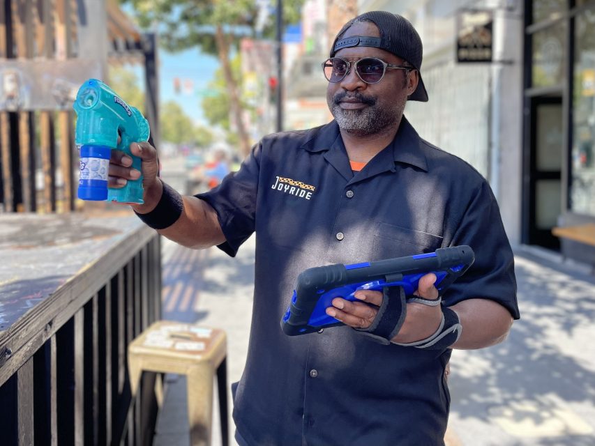 Man in a black shirt and cap holds a blue spray bottle and a tablet while smiling on a sunny street.