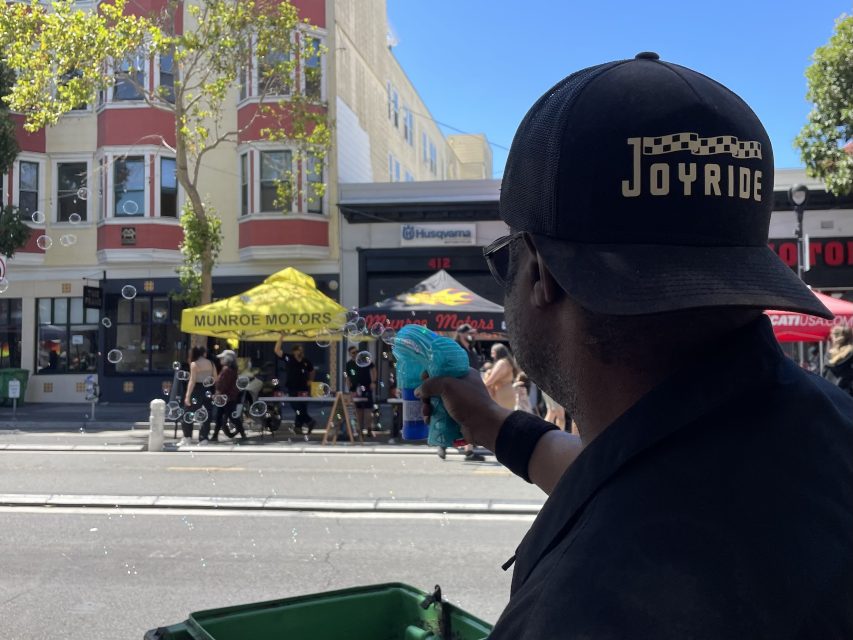 A person in a Joyride hat uses a toy to blow bubbles on a street with shops and people in the background.