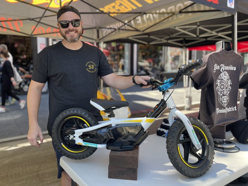 A man in sunglasses and a black t-shirt stands beside a small electric dirt bike on a table under a tent at an outdoor event. Shirts and other merchandise are also displayed.