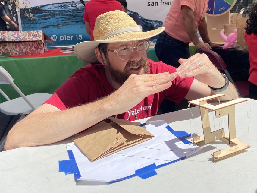 A man wearing a straw hat and glasses is demonstrating a string mechanism on a wooden device at an outdoor event. A brochure and various crafts are on the table.