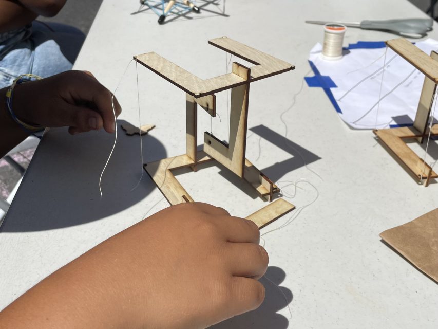 A close-up of hands assembling a wooden structure with string at an outdoor table. Scissors and other materials are visible in the background.