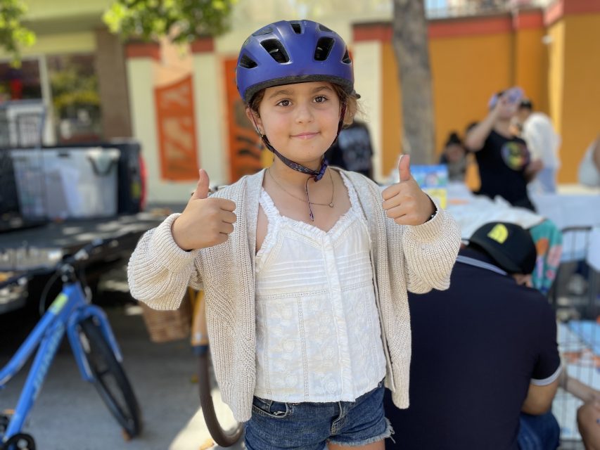A young child wearing a purple helmet, white top, and beige cardigan gives two thumbs up while standing outdoors. Bicycles and people are visible in the background.