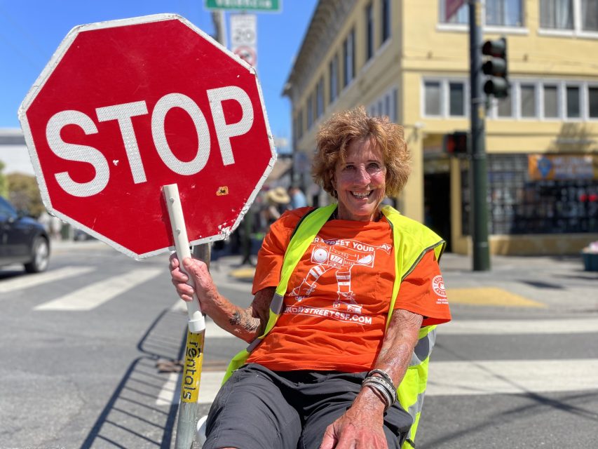 A person wearing an orange shirt and yellow vest holds a large stop sign while sitting in the street with a building and traffic lights in the background.