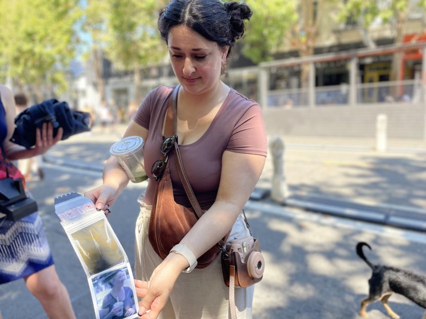 A woman wearing a brown top and beige pants shows a package with photos on a sunny sidewalk. She carries a tan handbag and a camera, and a can is attached to her bag strap.