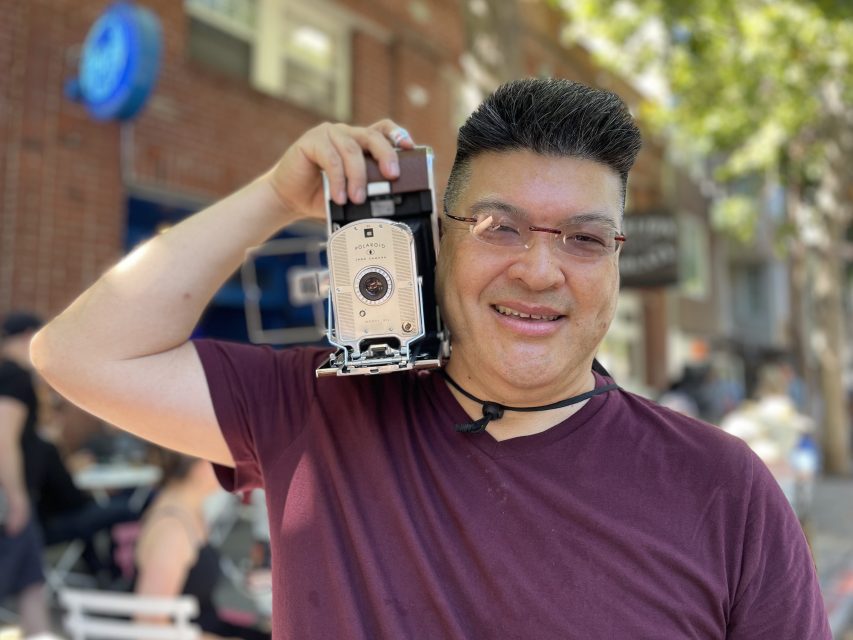 A person in a maroon shirt holds up an old-fashioned camera outdoors on a tree-lined street, smiling at the camera.