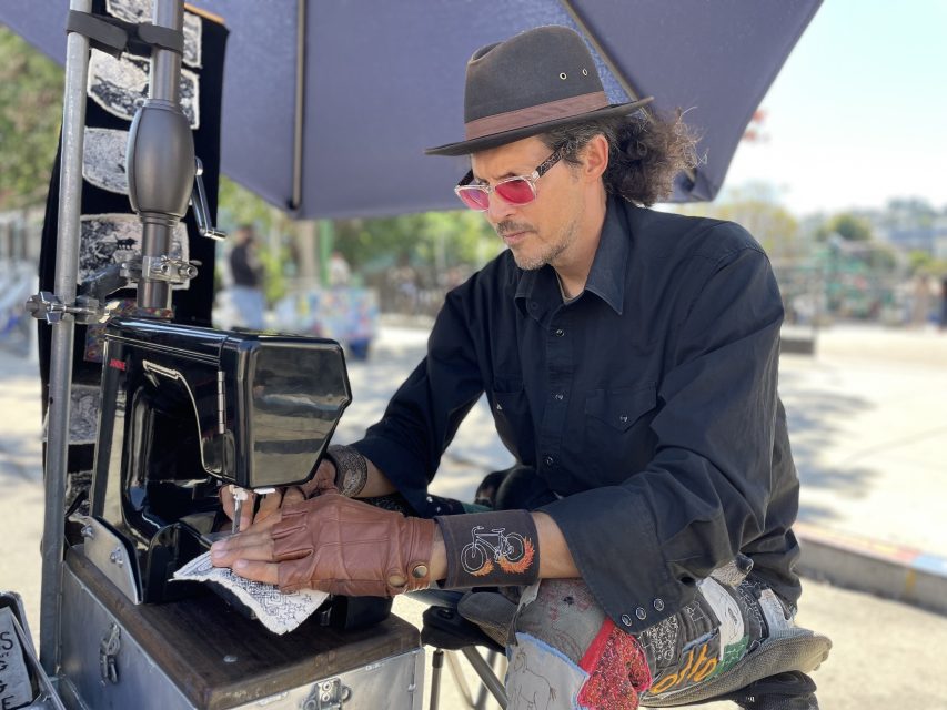 A person wearing a hat and sunglasses operates a sewing machine outdoors, underneath an umbrella. They are dressed in dark clothing with gloves.