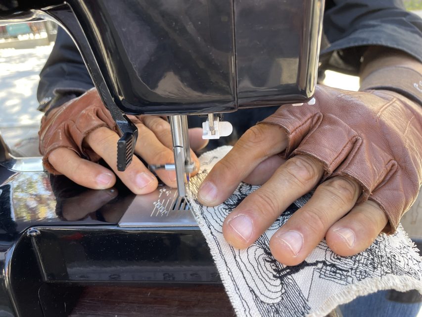 A person wearing fingerless gloves operates a sewing machine while stitching a piece of fabric with a patterned design.