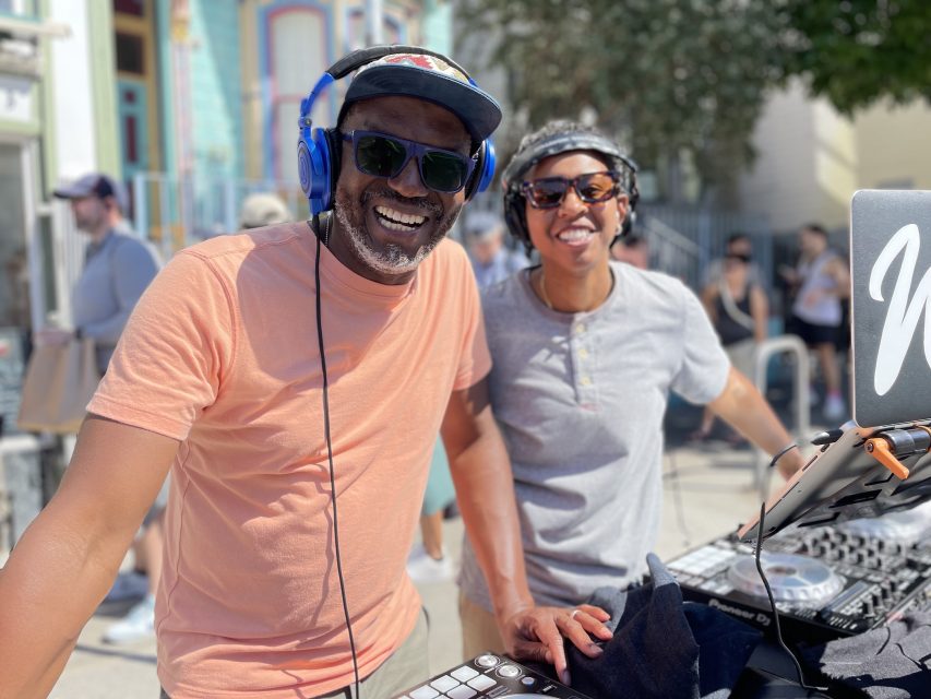 Two people are standing outside at a DJ booth; one wears a peach T-shirt, sunglasses, and blue headphones, and the other wears a gray shirt and sunglasses. Both are smiling.