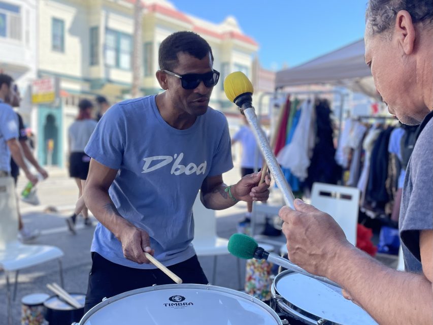 Two people are playing drums outdoors. One is holding drumsticks in each hand while wearing sunglasses and a blue shirt. The other, partially visible, is holding a mallet. They are performing at a street market.