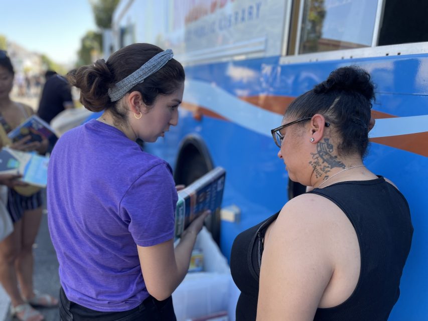 Two women are looking at books next to a blue library bus with an orange stripe. One woman is holding an open book, and the other is observing.