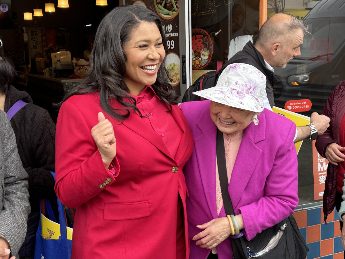 Two smiling women, one in a red blazer and the other in a pink jacket with a floral hat, share a joyful moment outside a restaurant. Other people and a storefront sign are visible in the background.