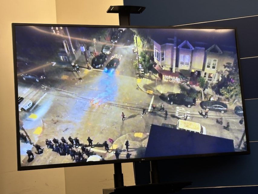 A large monitor displays a nighttime aerial view of a street intersection with numerous police officers gathering at the corner. Several police vehicles are present, and houses line the streets.