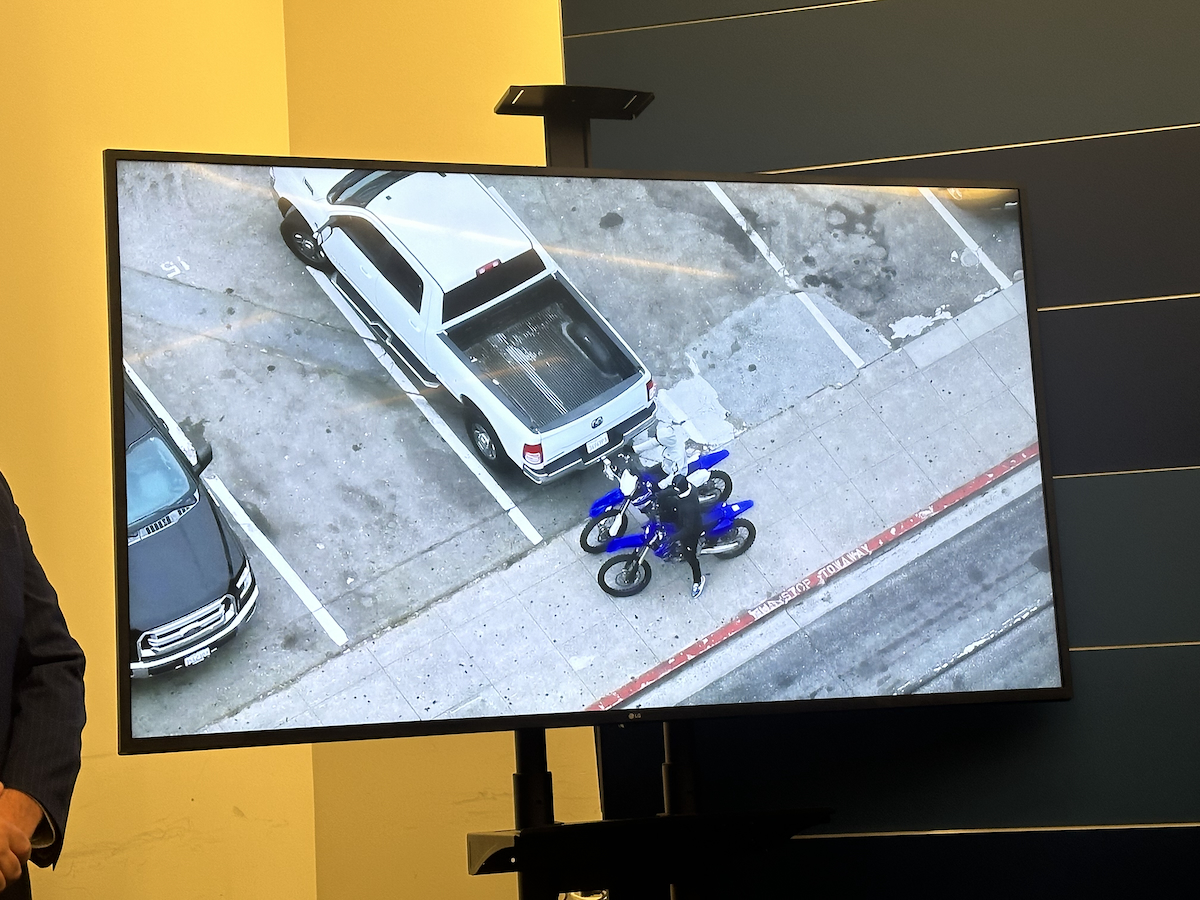 A television screen displays an aerial view of a white truck parked in a lot, with a blue motorcycle and a person next to it.