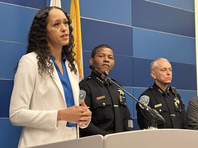 Brooke Jenkins in a white suit speaks at a podium with two uniformed police officers standing behind her against a blue-tiled wall.