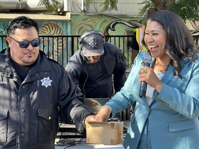 A man in a police uniform and Mayor London Breed in a blue blazer hold hands while the woman speaks into a microphone. A person in the background wearing a cap operates equipment.