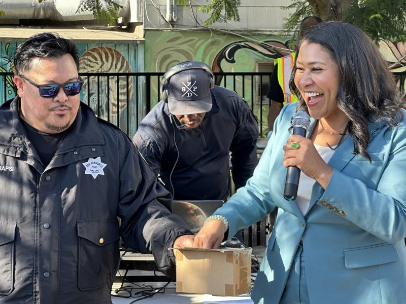 A man in a police uniform and Mayor London Breed in a blue blazer hold hands while the woman speaks into a microphone. A person in the background wearing a cap operates equipment.