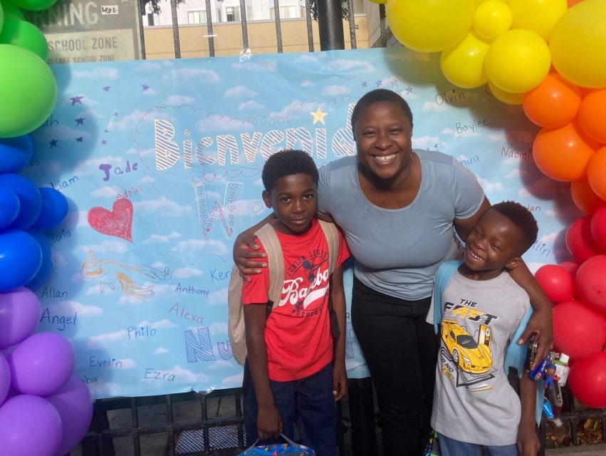 A woman stands smiling with two boys in front of a colorful balloon arch next to a "Back to School" sign. The boys are holding backpacks and smiling.