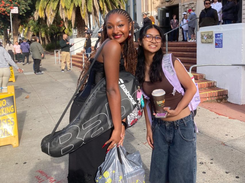 Two women smiling outdoors; one carries a tennis racket and shopping bags, the other holds a drink and snack. People and ornate building entrance in the background.