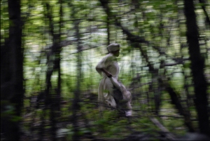 A person wearing white clothing runs through a blurry, densely wooded forest.