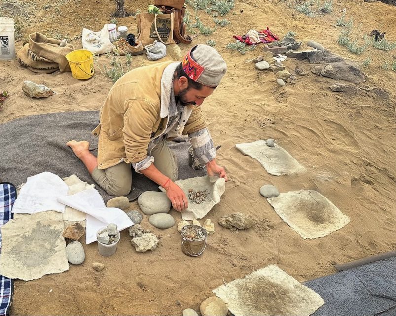 A person kneels on the ground working with various stone tools and pieces of cloth spread out around them, likely engaging in an archaeological or crafting activity in a sandy outdoor setting.