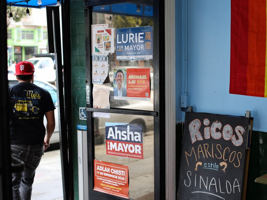 一扇門上貼有各種政治競選海報，右側有手寫的黑板廣告 Ricos Mariscos Estilo Sinaloa。左側有一個戴紅帽的人走過。