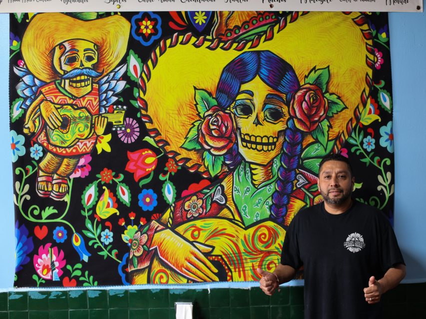 一位男士站在一幅色彩豐富的壁畫前，壁畫描繪了帶有花卉和心形圖案的骷髏，其中一個骷髏戴著黃色闊邊帽彈吉他，另一個有辮子髮型和玫瑰花。