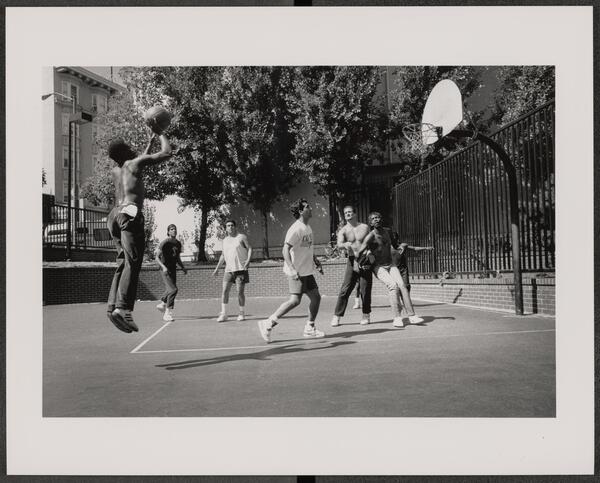 A group of men play a game of basketball on an outdoor court; one player is airborne near the hoop, holding the ball, while others watch or move around the court. Trees and buildings are in the background.