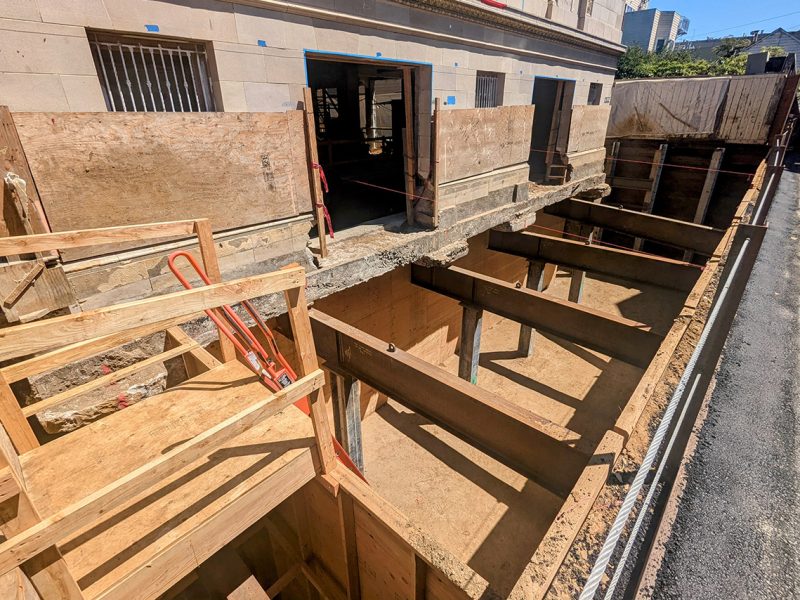 Construction site showing a partially excavated area with wooden support beams, scaffolding, and an exposed section of a concrete building.