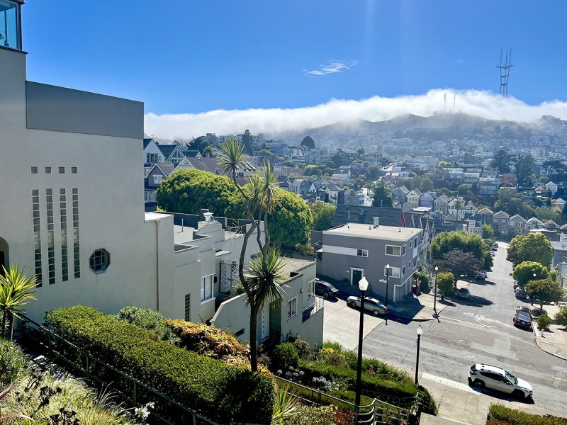 A fog-covered hill with a tower overlooks a neighborhood with modern houses and green trees under a clear blue sky.