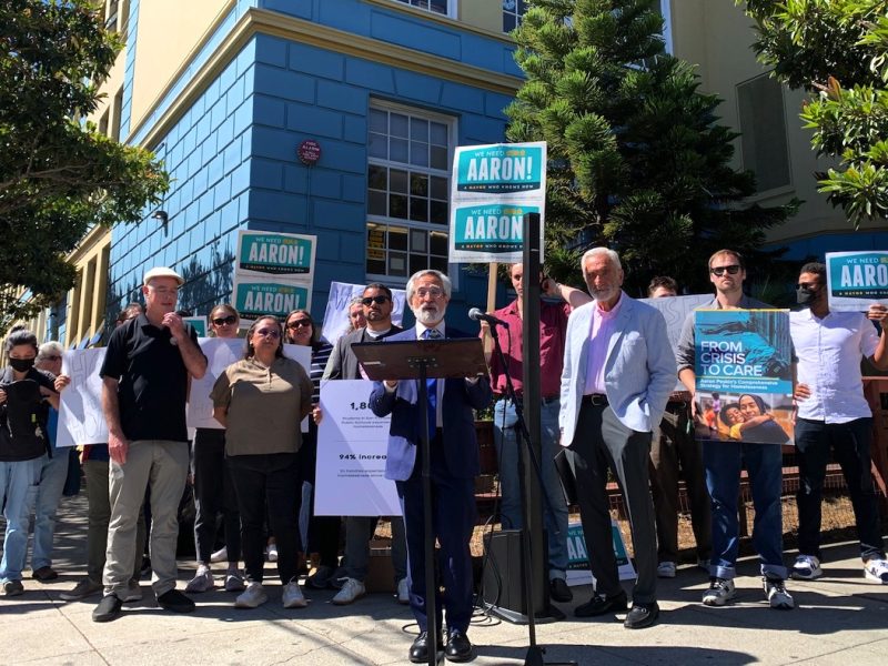 一群人站在一棟建築物外，舉著寫著「亞倫！」的牌子。和一張大海報。一個穿著西裝的人在講台上對大家說話。