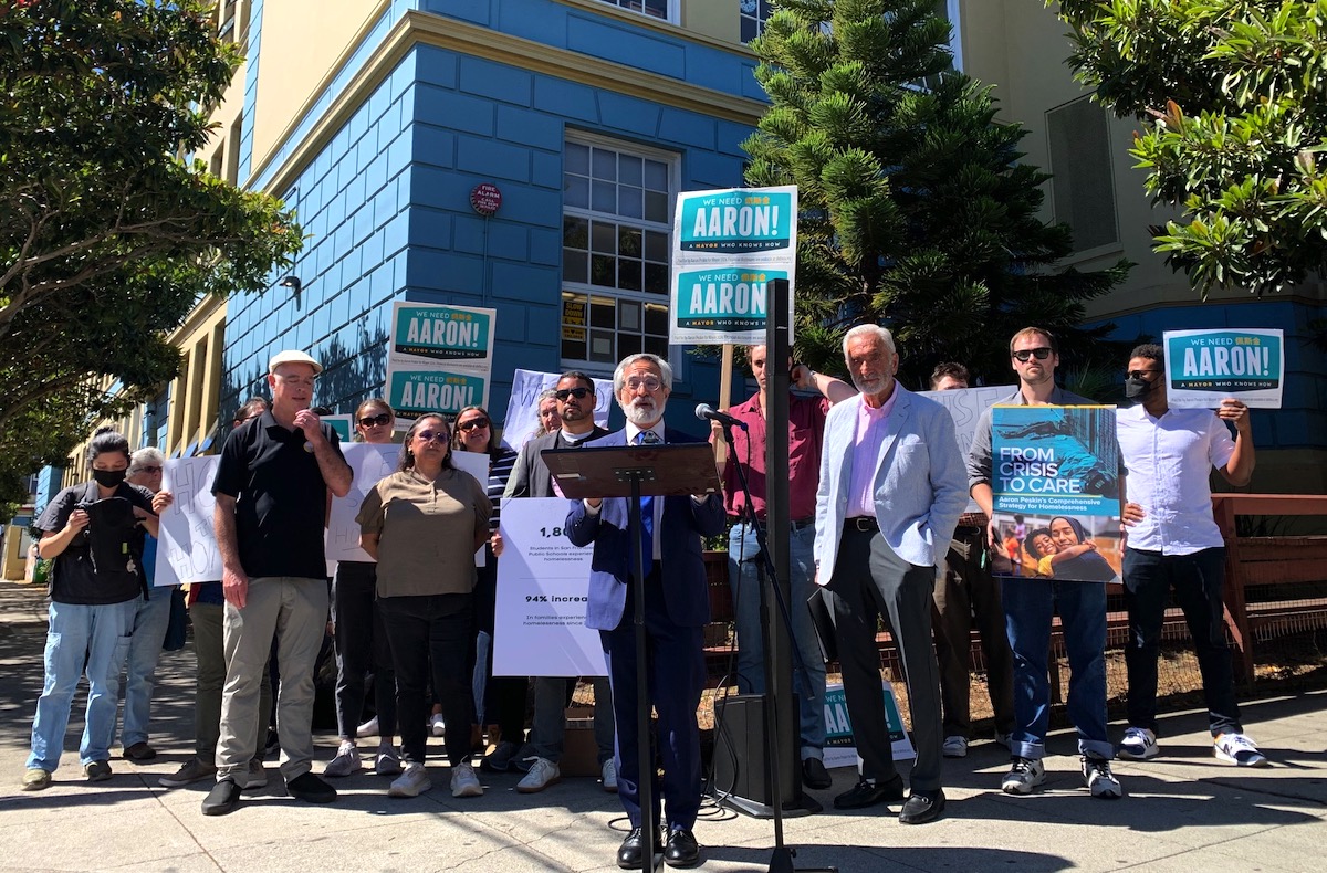 一群人站在一棟建築物外，舉著寫著「亞倫！」的牌子。和一張大海報。一個穿著西裝的人在講台上對大家說話。