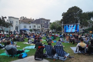 People gathered on a lawn, sitting on blankets and chairs, watching a large outdoor screen in a park. Houses and trees are visible in the background.