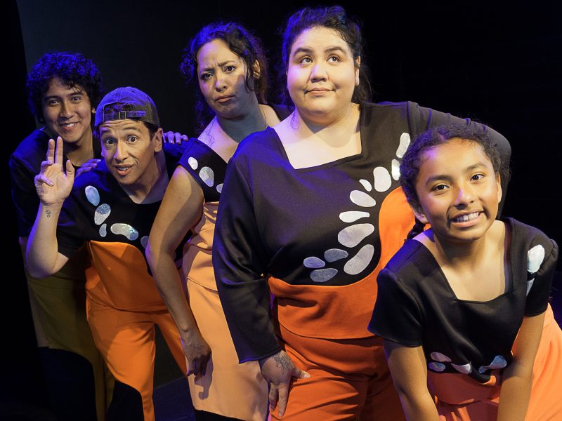 a group of five Latinx youths in orange bottosm and black and white tops pose while in a play on stage for Fantasmitas