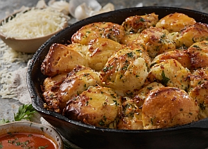 A skillet filled with golden-brown garlic butter smashed potatoes garnished with herbs. In the background, there is a bowl of shredded cheese and a small dish of sauce.