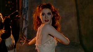 A woman with red hair, dressed in a white garment, looks over her shoulder with an expression of surprise or fear in a dimly lit environment.