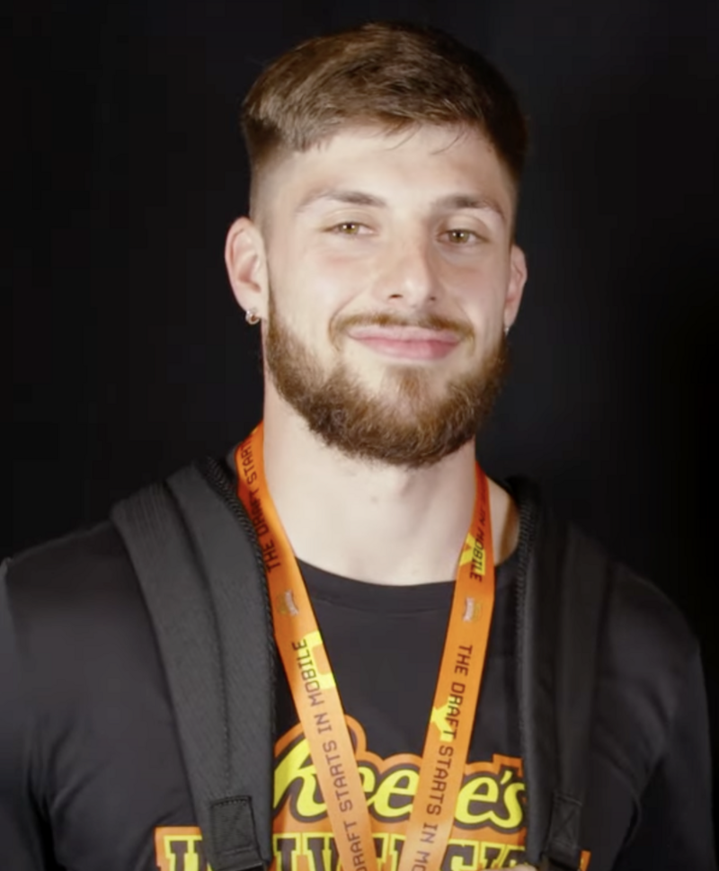 A man with a beard and short hair smiles at the camera, wearing an orange lanyard and a black backpack.