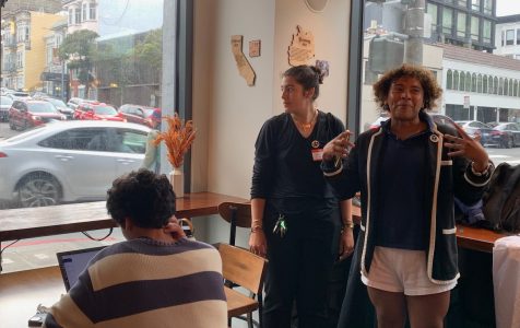 Three individuals are indoors near a large window. One is sitting and using a laptop, another stands and looks outside, and the third is gesturing and facing the camera.