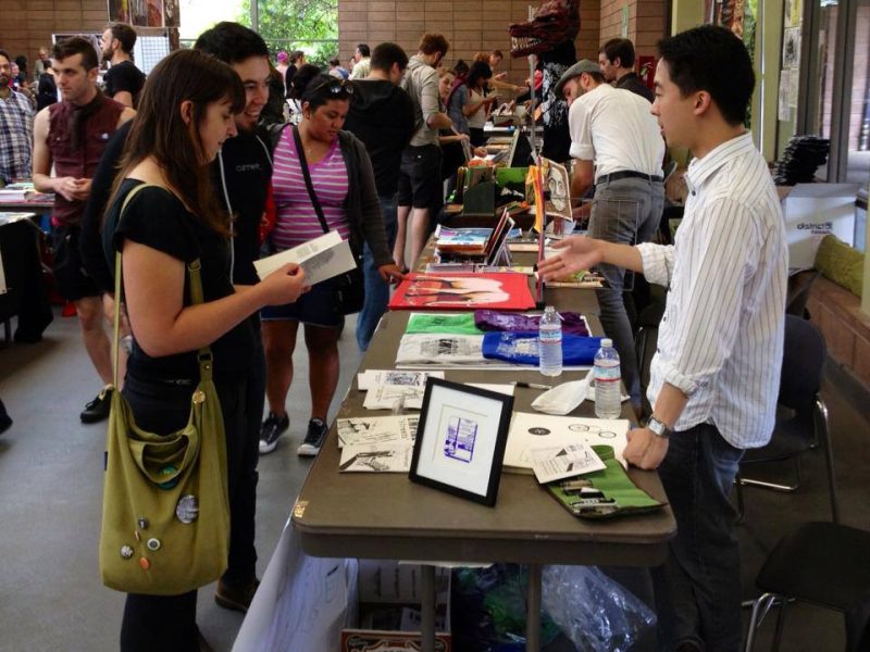 People at a convention booth engaging with a vendor displaying various art pieces and printed materials. Several attendees are browsing the items while others walk through the room.
