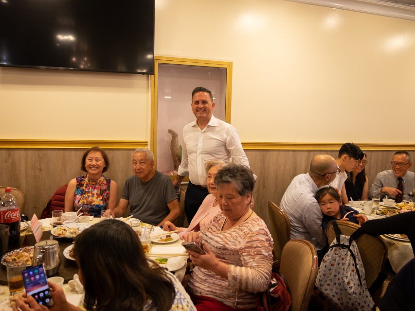 A group of people gathered around a table in a restaurant, with one person standing while others are seated, eating, and using their phones.