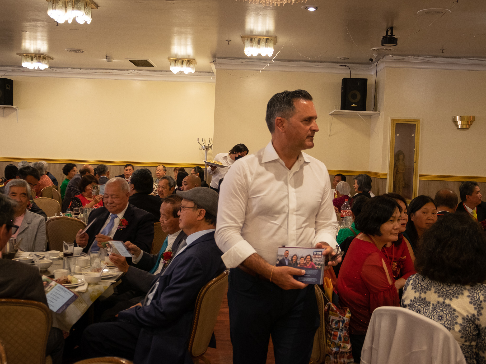 A man in a white shirt stands holding pamphlets in a room filled with seated people attending an event.