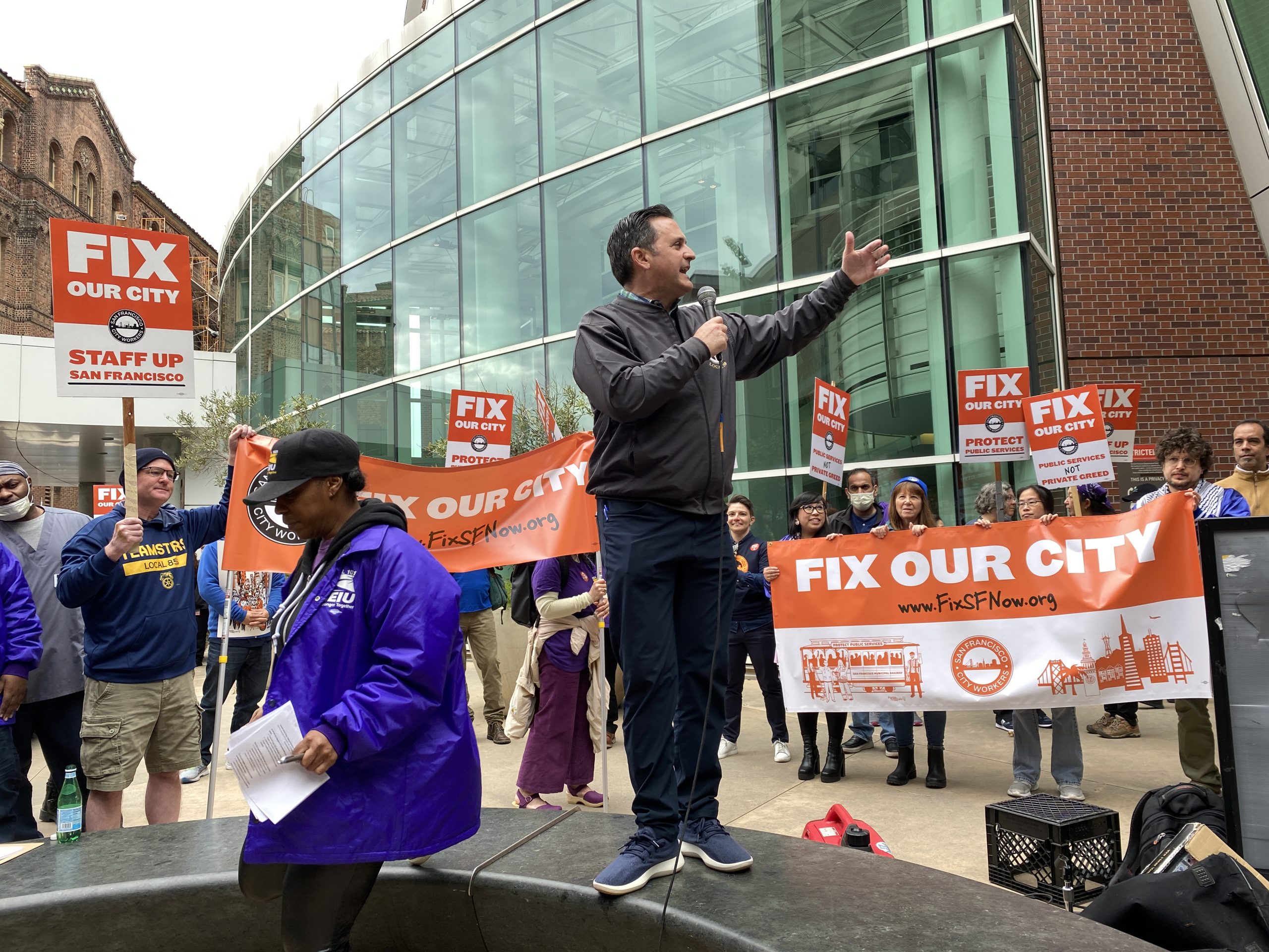 A man speaks to a crowd at a rally with signs that read "FIX OUR CITY." People hold banners and signs, and the event appears to be taking place in an urban area with modern buildings in the background.