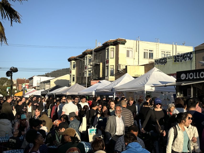 A crowded street market with people browsing booths and stalls under white canopies on a sunny day, with buildings in the background.