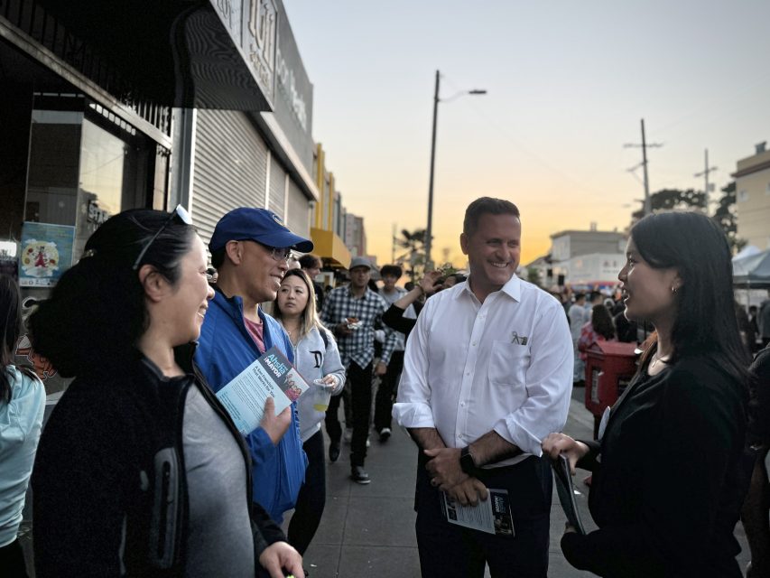 A group of people converse on a street during an event. One person holds a brochure, and a man in a white shirt smiles while engaging in the discussion. Others and booths are visible in the background.