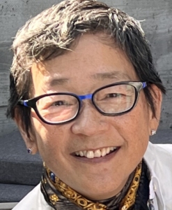 A person with short hair and glasses smiles at the camera. They are wearing a patterned scarf and a light shirt.