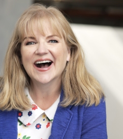 A woman with shoulder-length blonde hair is smiling brightly. She is wearing a blue cardigan over a white shirt with colorful flower patterns.