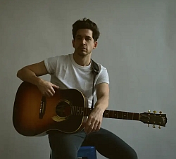 A man in a white t-shirt is sitting on a blue stool and holding an acoustic guitar against a plain background.