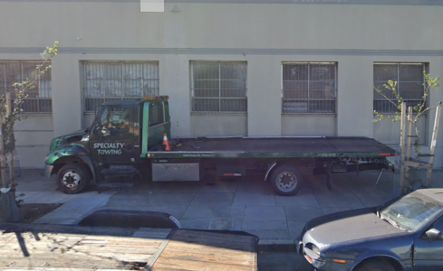 A green specialty towing truck is parked on the street, next to a beige building with barred windows.
