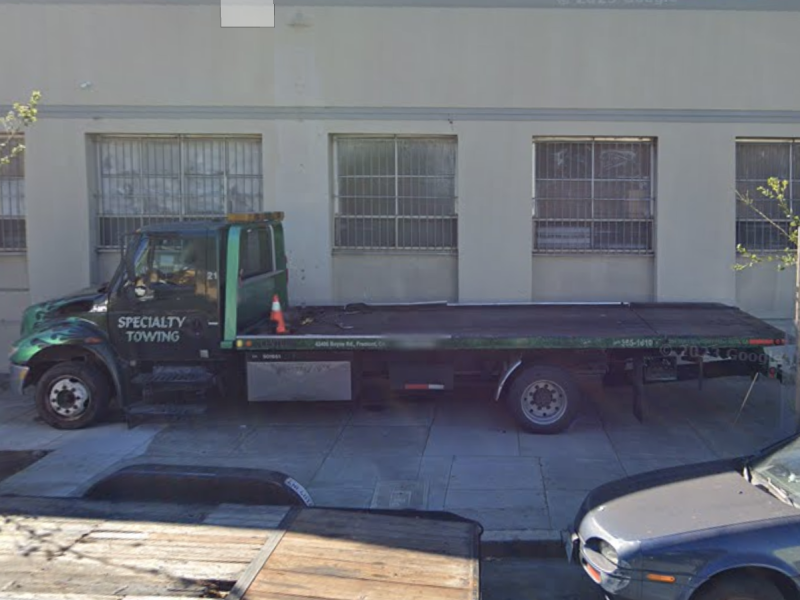 A green specialty towing truck is parked on the street, next to a beige building with barred windows.